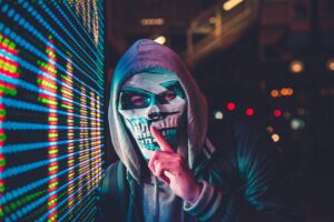 フードを被りマスクで顔を隠したハッカーが左手の人差し指を口に当て、静かにしろという合図を送っている様子